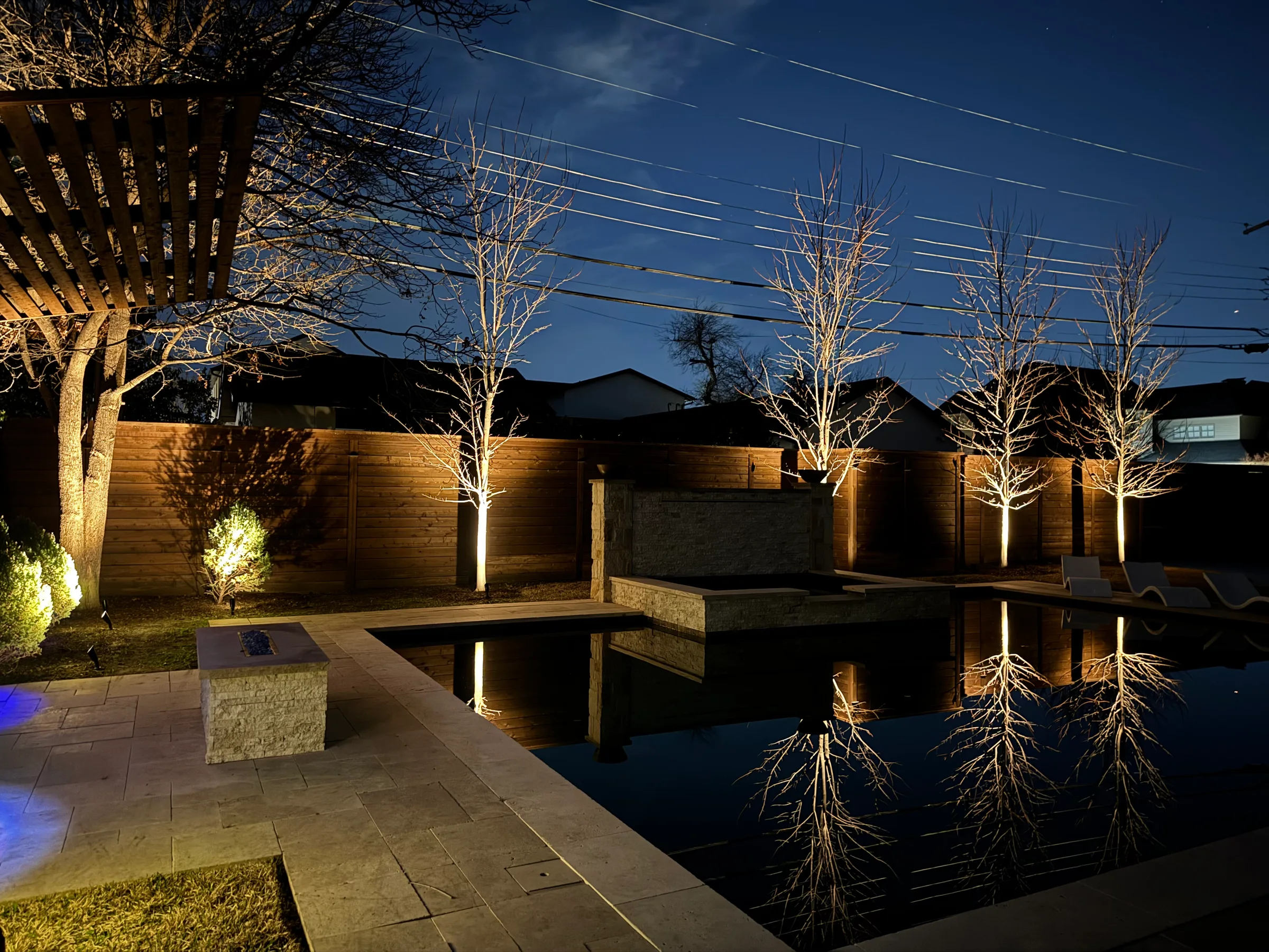 Pool terrace, trees, and wood privacy wall illuminated with warm landscape lighting
