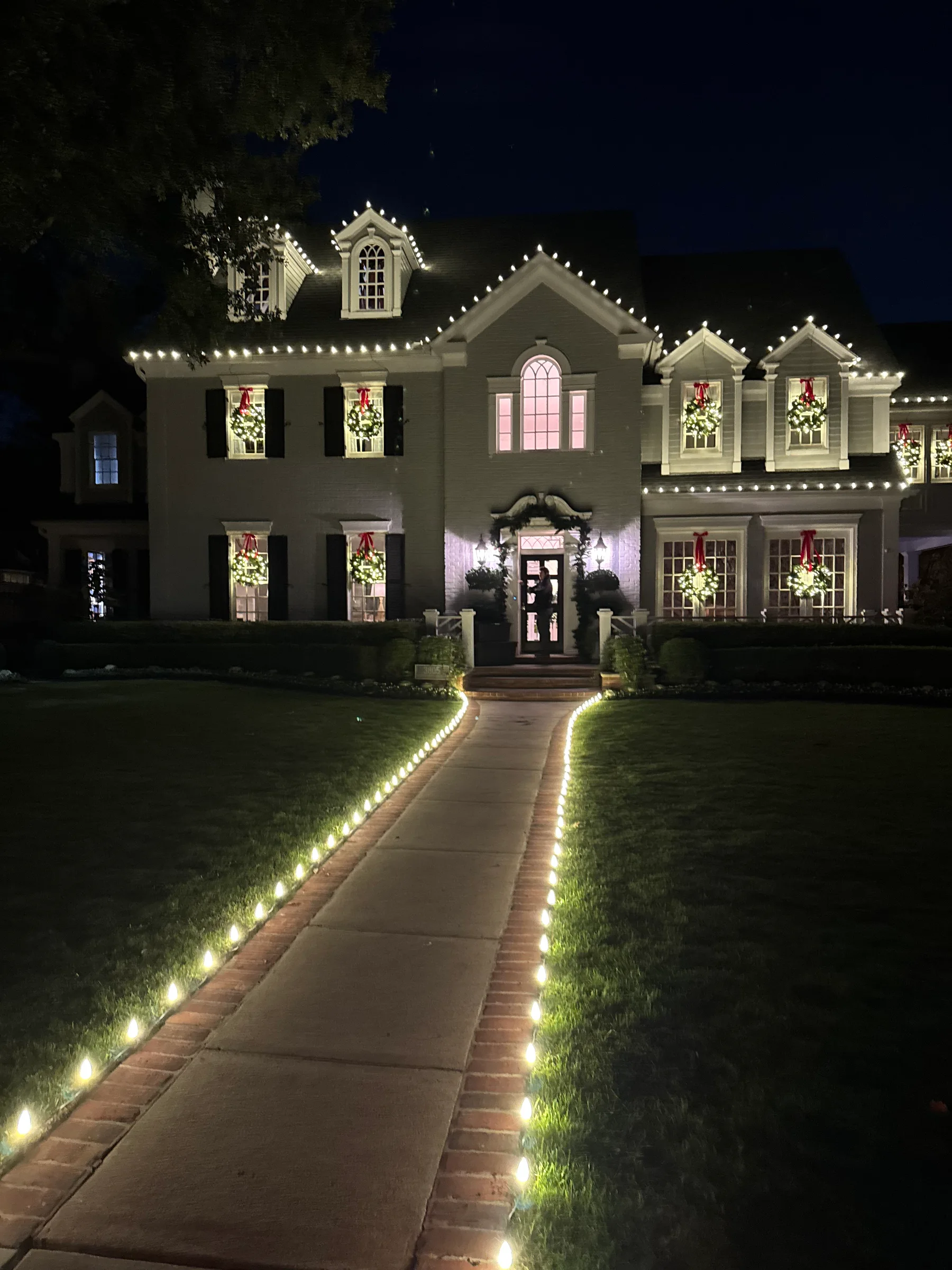 Gray two-story home with warm white roofline lights, window wreaths, and an illuminated front walk