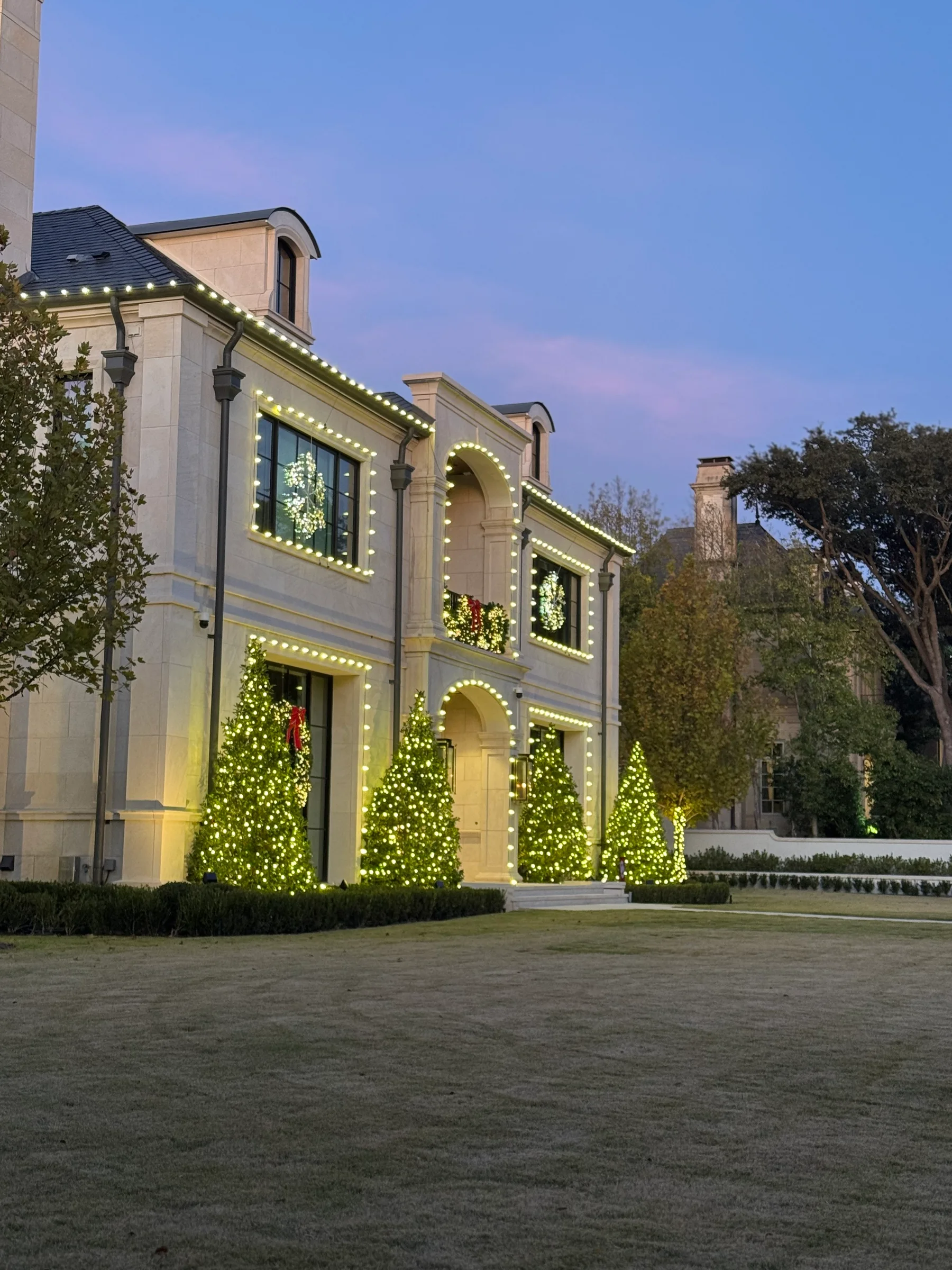 Large light-colored home outlined with warm white Christmas bulbs around roof peaks, windows, and arches