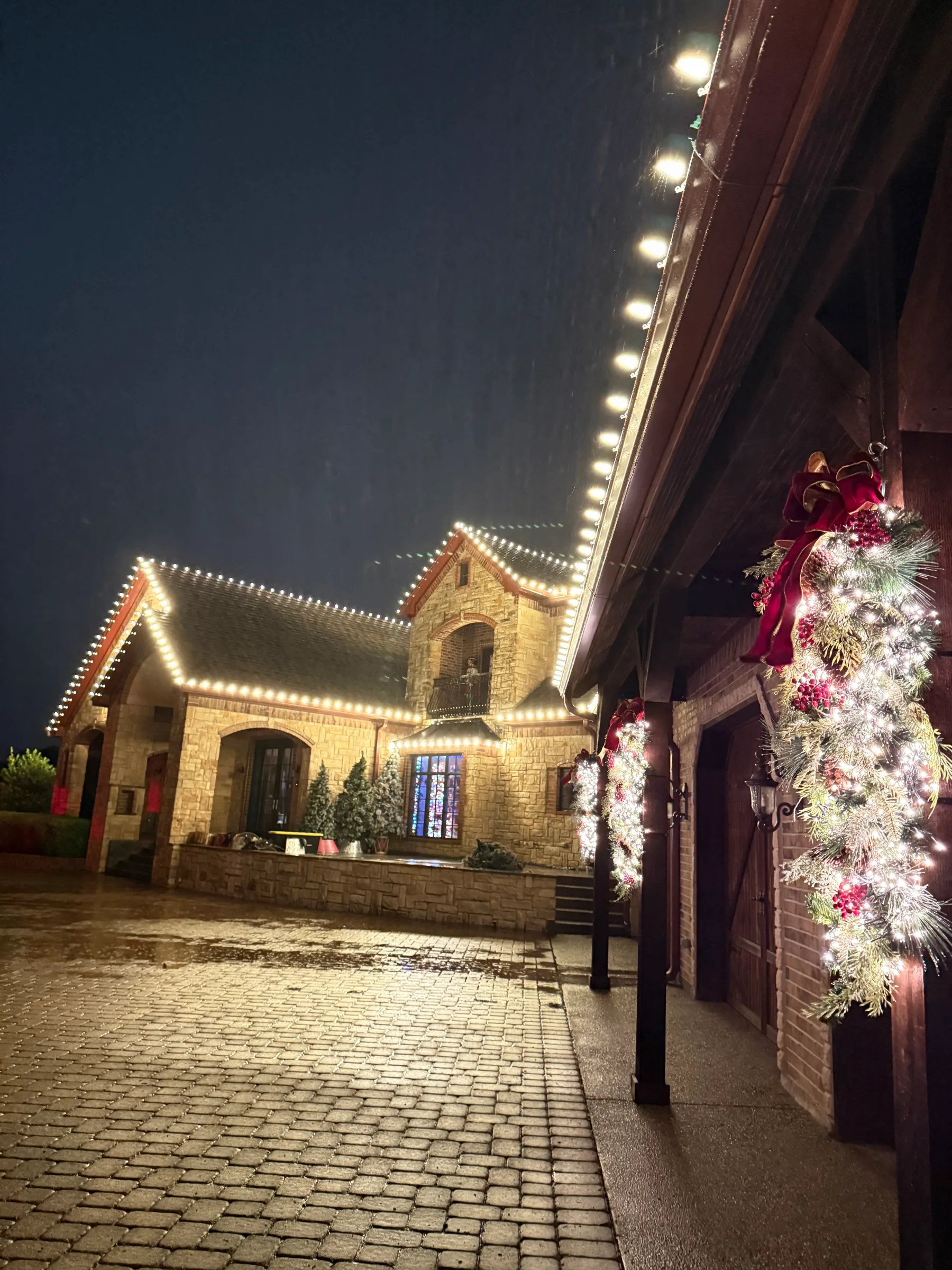 Stone courtyard with warm white roofline lights and lit Christmas garland on porch columns